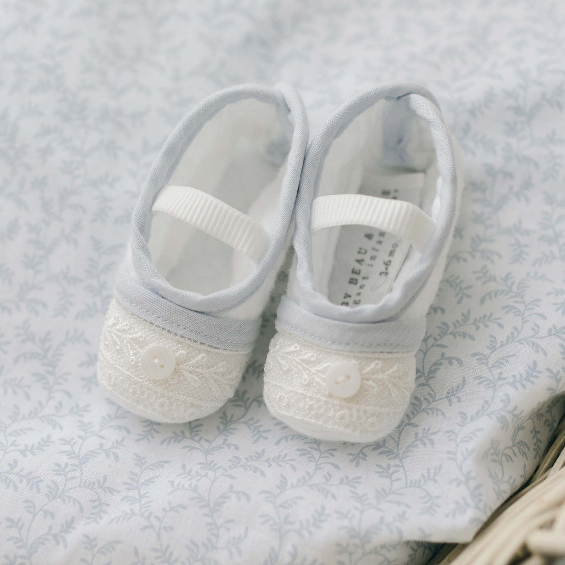 The Harrison Booties, a pair of baby shoes handmade in the USA, feature white lace and button details on the toes, with gray fabric and white straps across the top. They are elegantly displayed on a light blue patterned cloth and make a perfect heirloom gift.