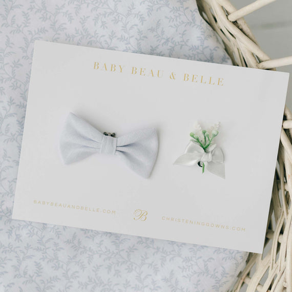 A flat lay display of a pale blue linen bow tie and a delicate boutonniere with white ribbon and green accents, showcased on a branded Baby Beau & Belle card.