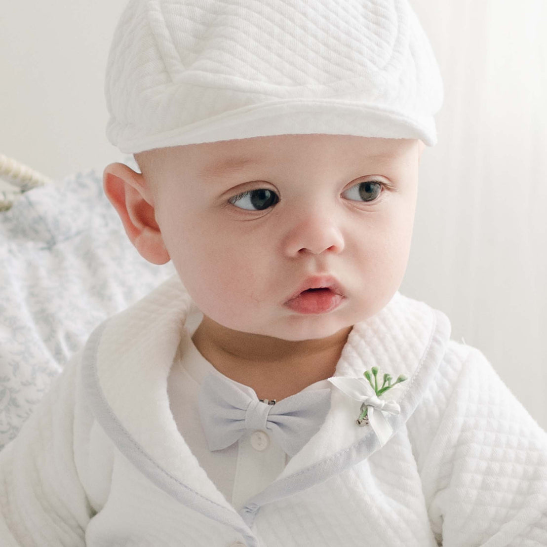 A baby dressed in the Harrison Linen Bow Tie & Boutonniere set—featuring a white textured outfit, matching cap, small bow tie, and decorative boutonniere on their jacket. The baby, clad in soft blue linen/cotton fabric, has a curious expression and gazes slightly to the side. The softly blurred background accentuates the baby's cuteness.