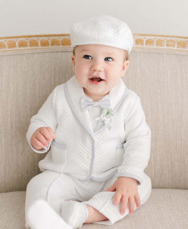 A baby in a white quilted outfit and matching hat sits on a beige sofa. The baby smiles while looking slightly upward, wearing a bow tie and a small boutonniere. The sofa has a wooden frame, and the background is a neutral wall.
