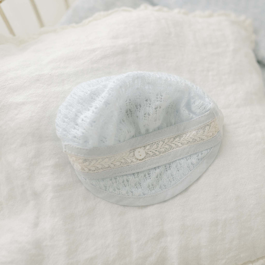 The Harrison Blue Knit Hat, handmade in the USA, rests gently on a soft, white pillow. Adorned with intricate lace trim and a small button at its center, it exudes vintage charm. The background of lighter blue fabric enhances the newborn baby's comfort.