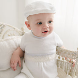 Baby wearing the Harrison Short Sleeve Romper with quilted fabric and lace detailing, paired with a matching white quilted newsboy cap, seated in a vintage wicker crib.