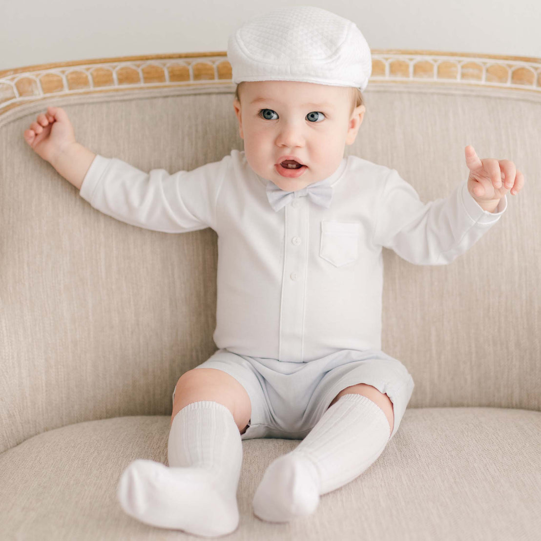 Baby boy sitting on a chair and wearing a white onesie with light blue shorts, a Harrison Quilted Newsboy Cap and blue linen bow tie 