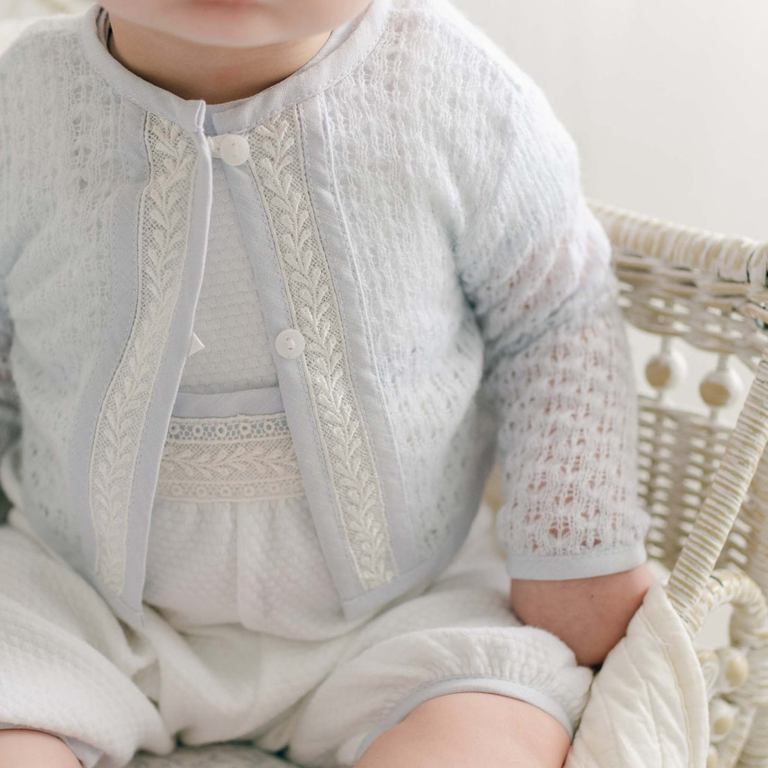 A baby wearing the Harrison Blue Knit Sweater over a white onesie sits upright in a wicker basket. The basket features a subtle woven pattern. The baby's face is partially visible, but the focus is primarily on their clothing and the basket.