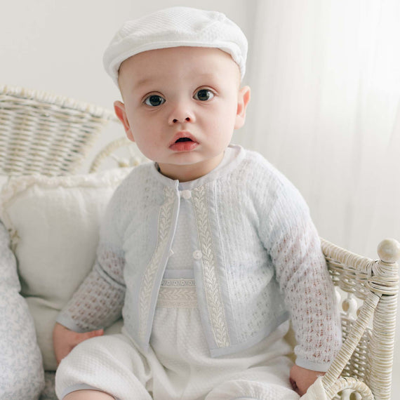 Baby boy sitting in a wicker cradle, dressed in a soft blue knit sweater with intricate lace details and white rompers.