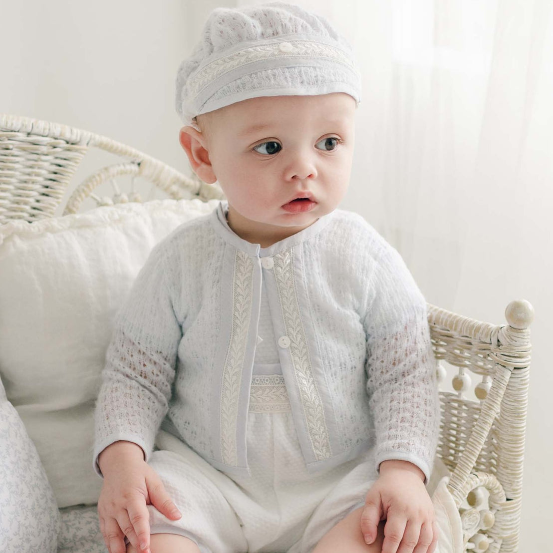 Baby boy seated on a wicker chair, dressed in a delicate blue knit sweater with ivory lace detailing and white quilted pants.
