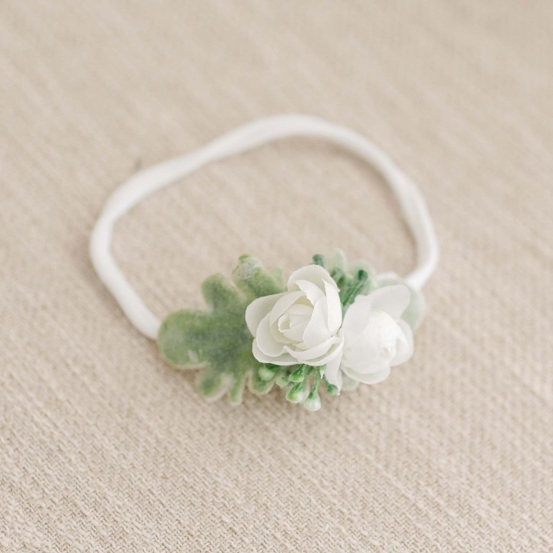 A delicate heirloom Eliza Flower headband featuring white flowers and green leaves on a soft beige fabric background.