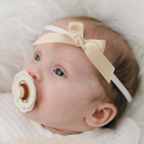 A close-up image of a baby with blue eyes, using a pacifier and wearing the Mia Bow Headband, a soft headband in ivory and detailed with a gold silk ribbon bow, lying on a fluffy white blanket.
