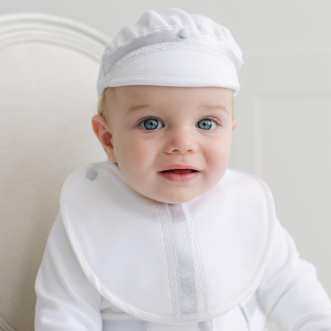 Baby in the Henry Bib and Brim Hat sitting on a beige chair. Details show blue silk ribbon with white Venice lace on the bib.