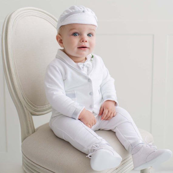 Baby in all white outfit sitting on a chair with a plain background