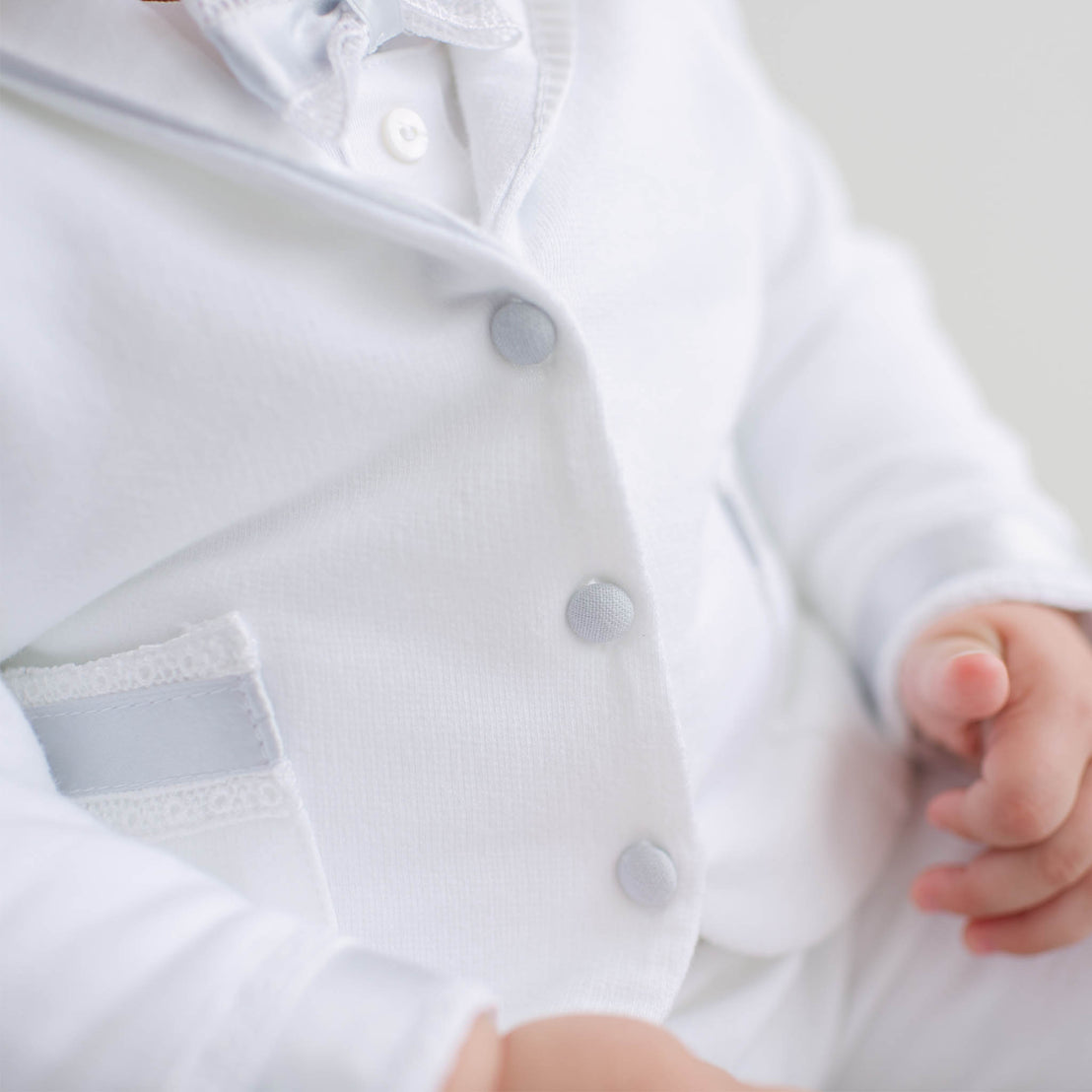 Close-up of a baby's white outfit with buttons and a hand holding a small object.