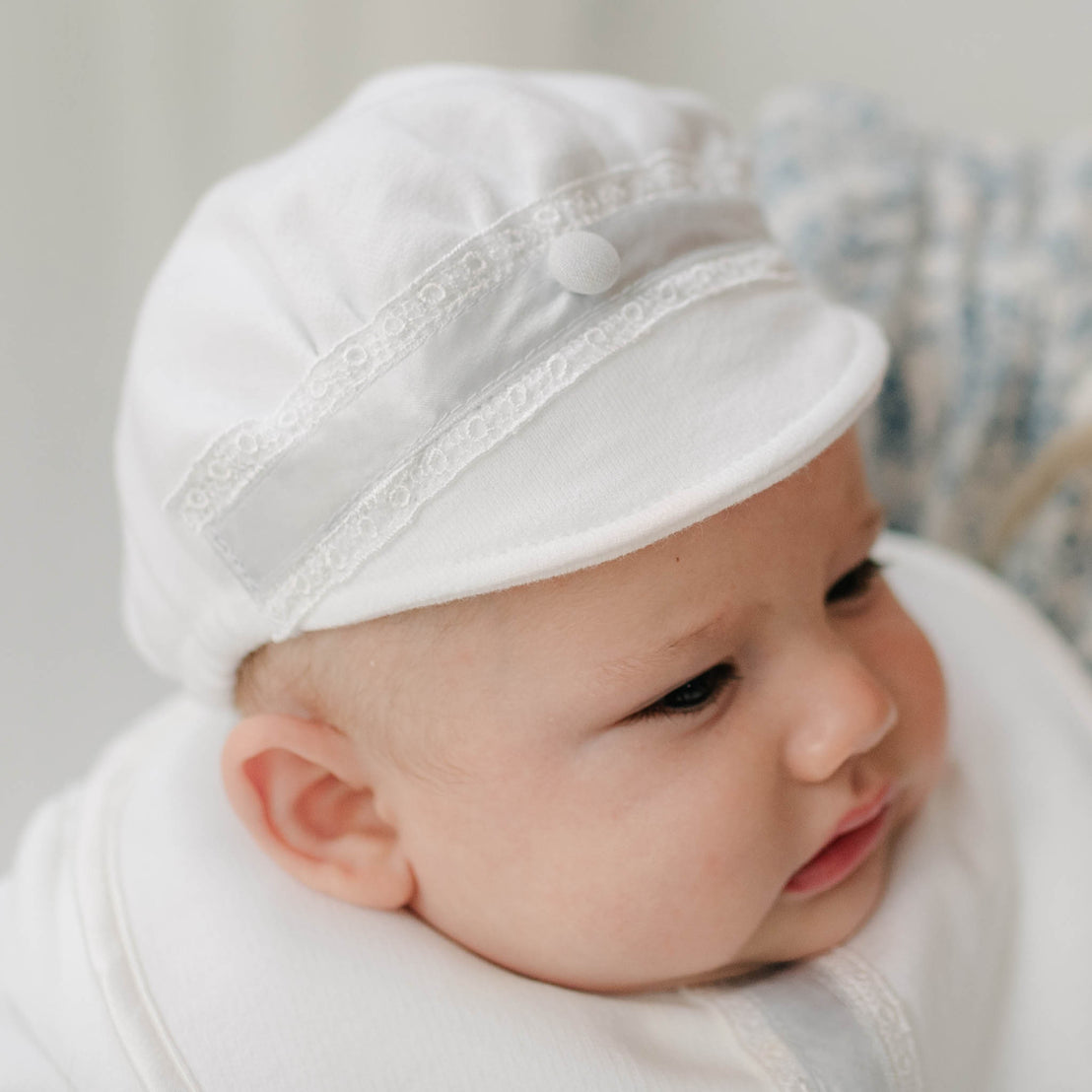 Close up of a baby wearing the Henry Brim Cap with lace details on a neutral background. The hat is crafted with white french terry and features Venice lace and blue silk ribbon with a blue linen button.