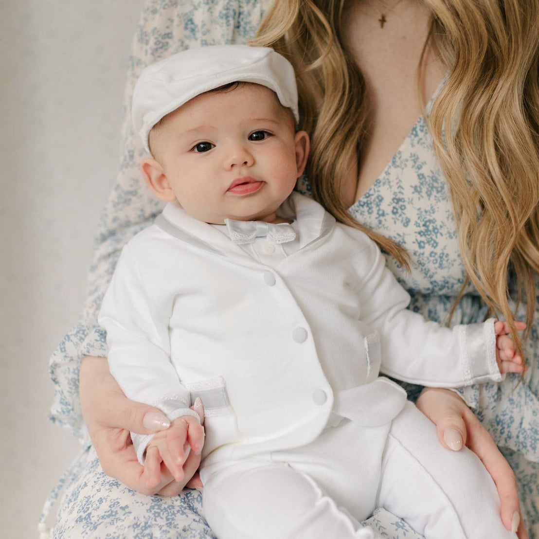 Baby in the Henry 3-Piece Suit and matching Brim Cap sitting on his mother's lap as she gently holds her hand. The jacket and bowtie features blue silk ribbon and lace detailing. 