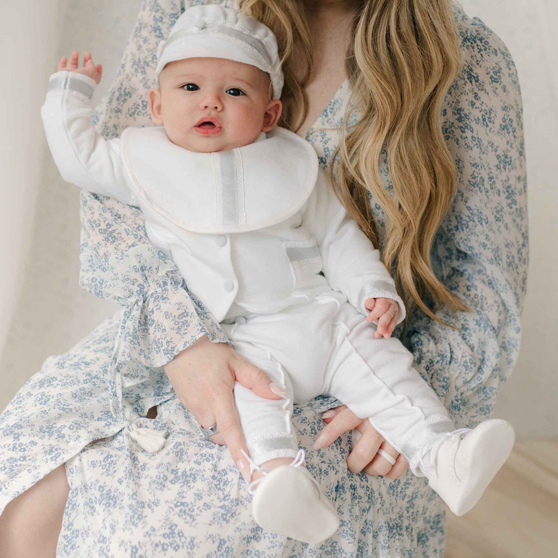 Close up of a baby in the Henry 3-Piece Suit being held by a woman wearing a floral dress. The suit features silk ribbon and Venice lace detailing along the cuffs and pockets. 