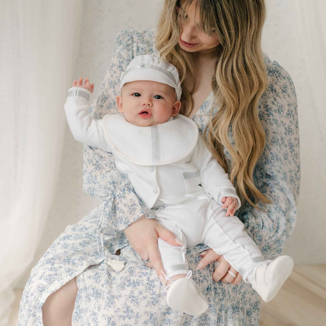 Woman in a blue floral dress holding a baby dressed in the Henry 3-Piece Suit and matching Bib. Set against a neutral background. Details on the bib and brim cap show Venice lace and silk ribbon detailing. Part of Baby Beau & Belle's Baby Boy Suit Collection.