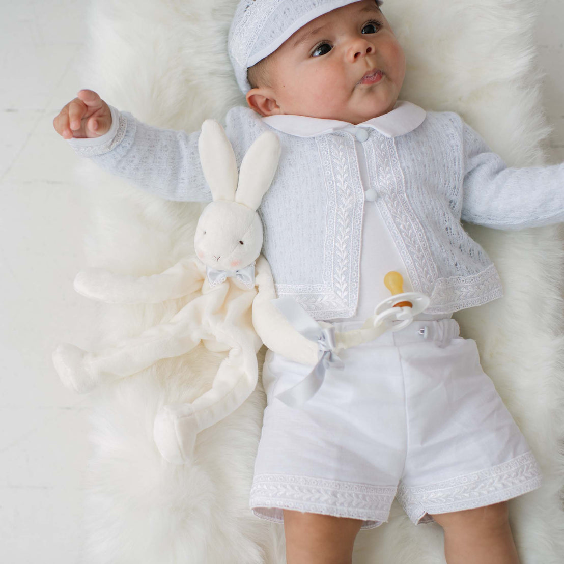 Baby in a blue knit sweater with matching shorts holding a plush bunny toy on a white background.