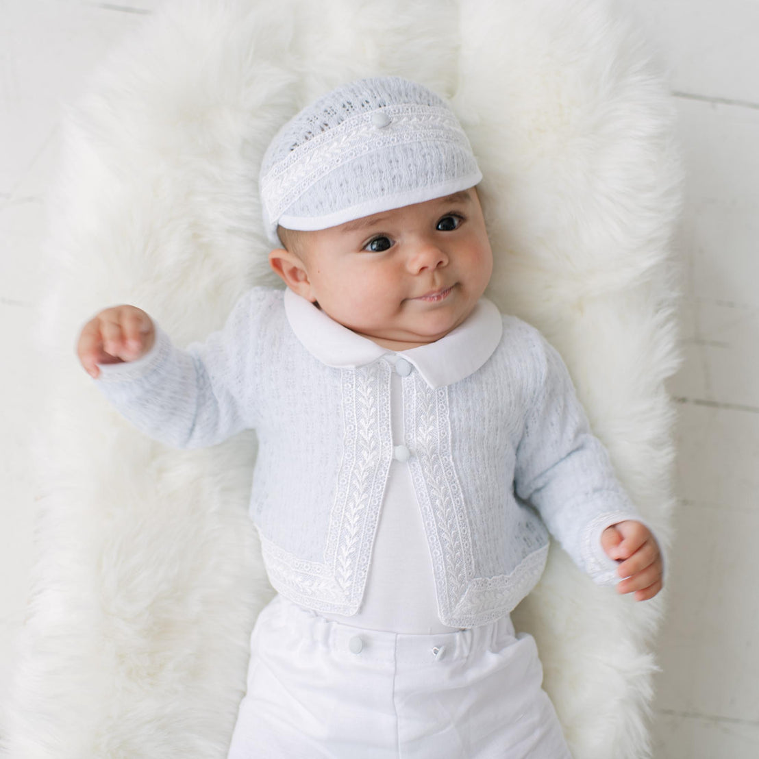 Baby wearing a white outfit and white knit sweater with matching hat on a white background.