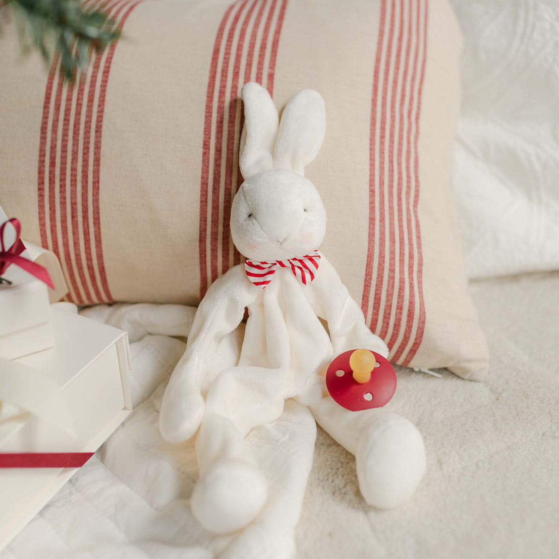 The Nicholas Silly Bunny Buddy Pacifier Holder, an ideal Christening gift, features a plush white bunny with a red and white striped bow around its neck. It rests on a beige pillow adorned with red stripes and holds a charming red pacifier. Accompanying this setup is a stack of elegantly wrapped white gift boxes tied with red ribbons.