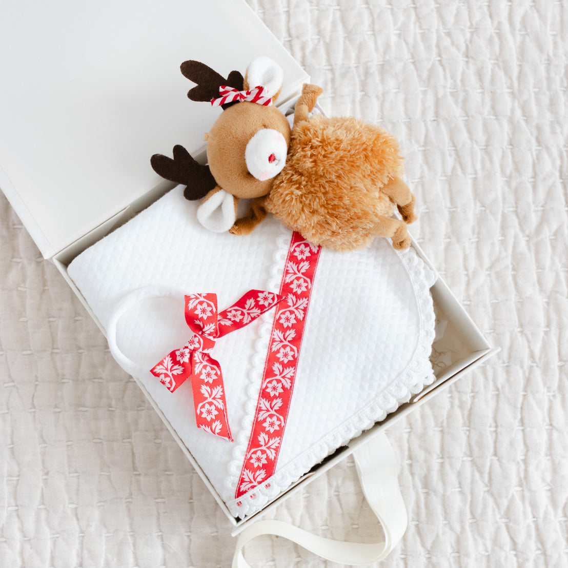 Plush reindeer toy with a decorative bow headband a top a matching white quilt blanket in a box.