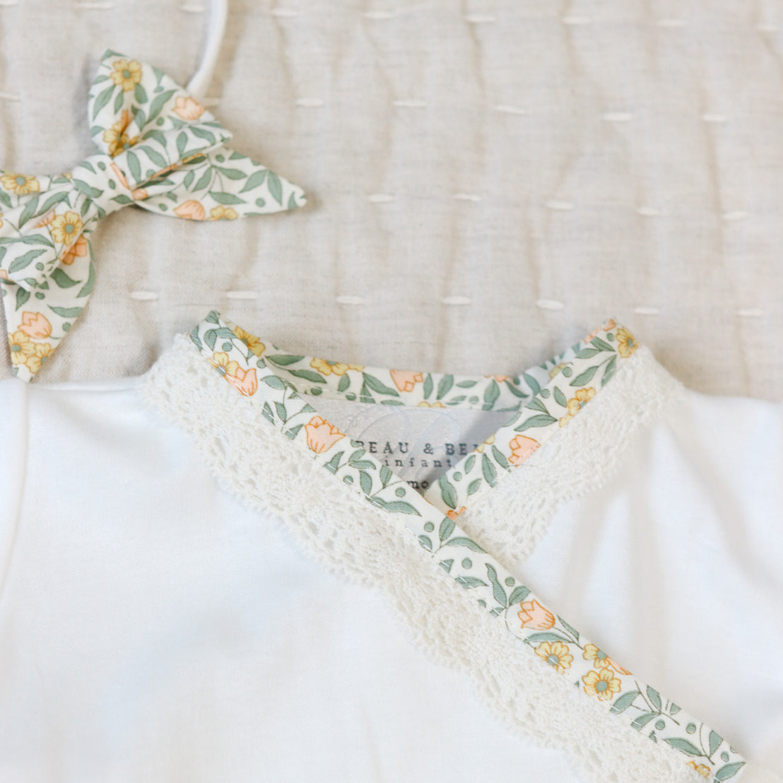 Close up flat lay photo of the Lillian Layette with yellow floral cotton and scalloped champagne lace trim. Set against a beige linen background.