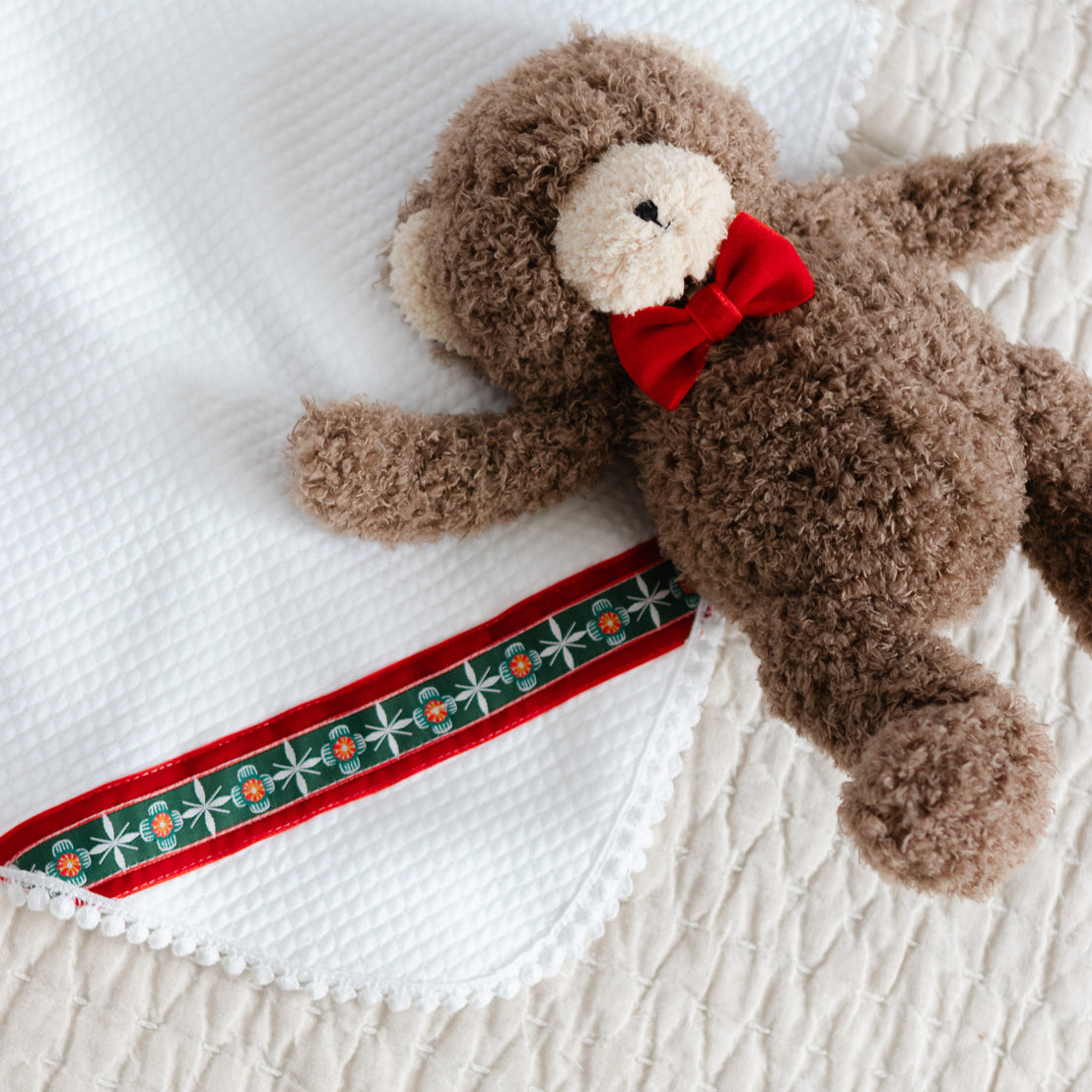 Brown teddy bear with a red bow tie lying on a white quilt blanket
