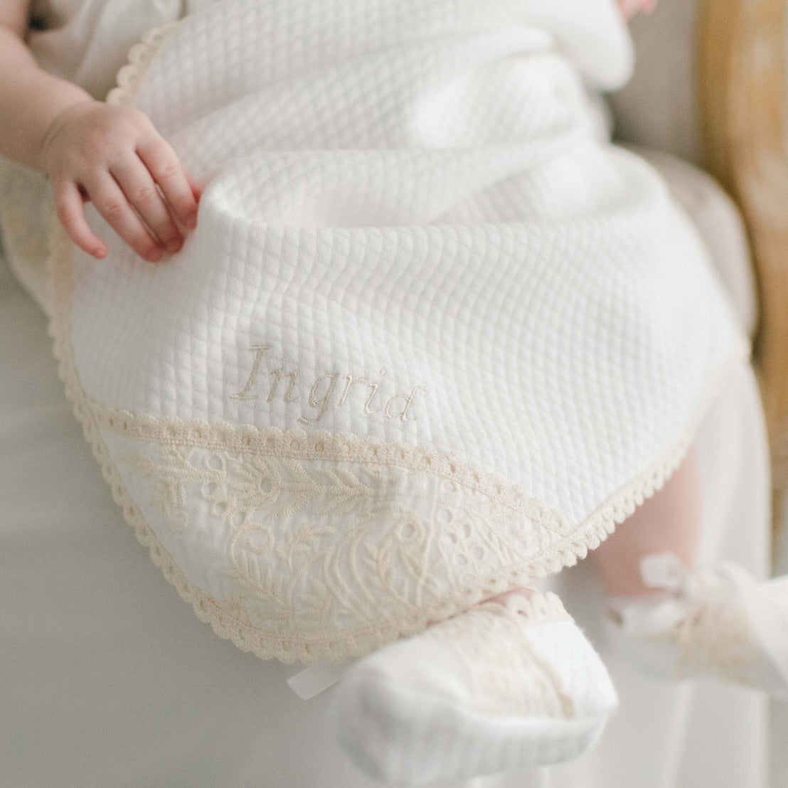 Baby covered in the Ingrid Blanket. The blanket is crafted with white quilted cotton and edged with a cotton lace in a natural champagne tone. The name "Ingrid" features above the lace corner in the same champagne thread