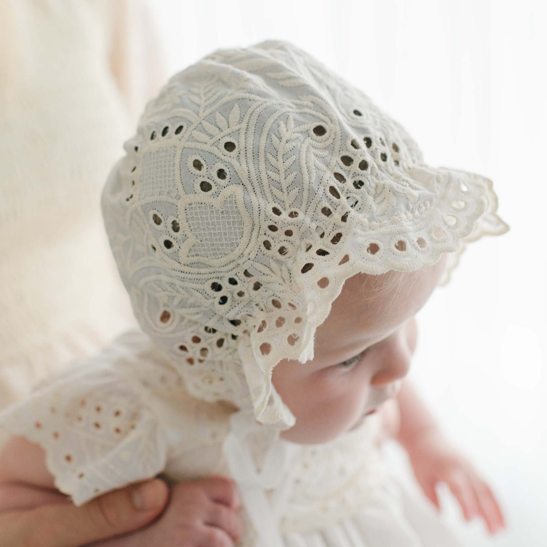 A close-up of a baby wearing an intricately patterned Christening Gown and the handmade in the USA Ingrid Bonnet. The baby is being gently supported by an unseen person. The delicate lace features detailed floral and leafy designs with scalloped edges, while the background is softly lit.