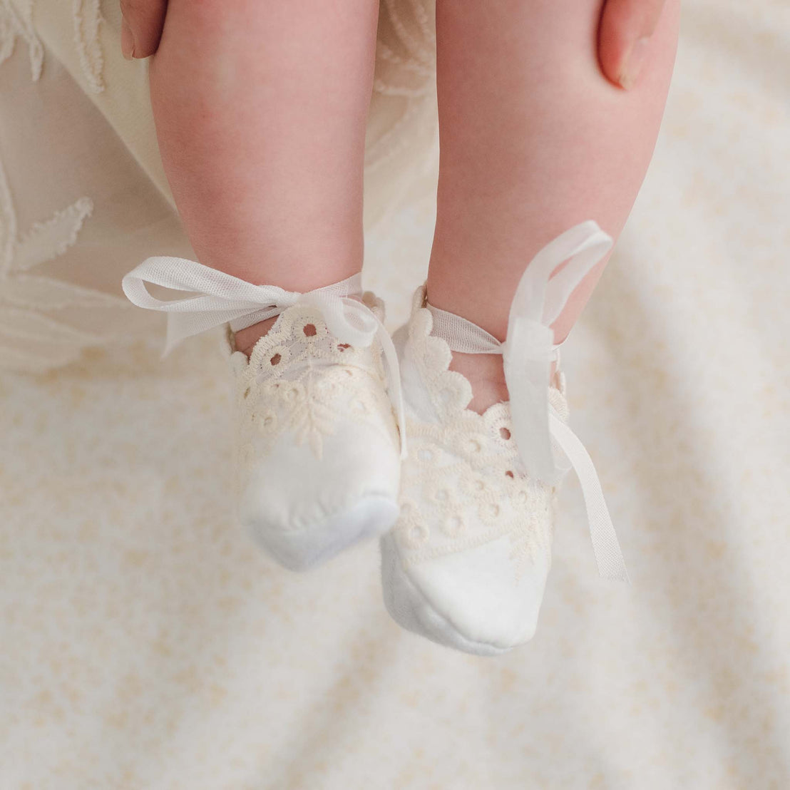Close-up of a baby's feet wearing delicate Ingrid Booties with vintage white lace and ribbons. The baby’s legs rest against a soft, light-colored fabric adorned with subtle floral embroidery. The booties feature intricate lace detailing, complementing the baby's fair skin perfectly.