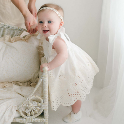 A smiling baby dressed in the Ingrid Romper Dress and matching headband is standing while holding onto a wicker chair, supported by an adult hand behind her. The scene showcases this charming girls' dress, which is handmade in the USA, and is illuminated by warm, natural light.