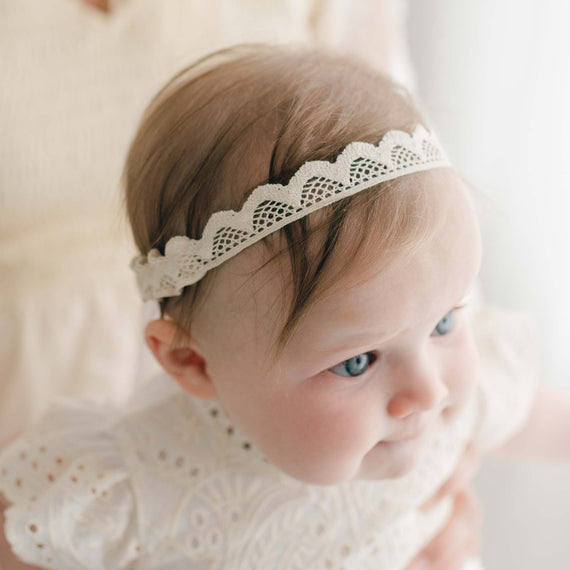 A close-up of a baby wearing an Ingrid Lace Baby Headband and a white lace outfit that resembles a christening gown. The baby, with light hair and blue eyes, looks slightly to the side while being held by an unseen person in a light-colored top. The background is softly lit, creating a gentle ambiance.