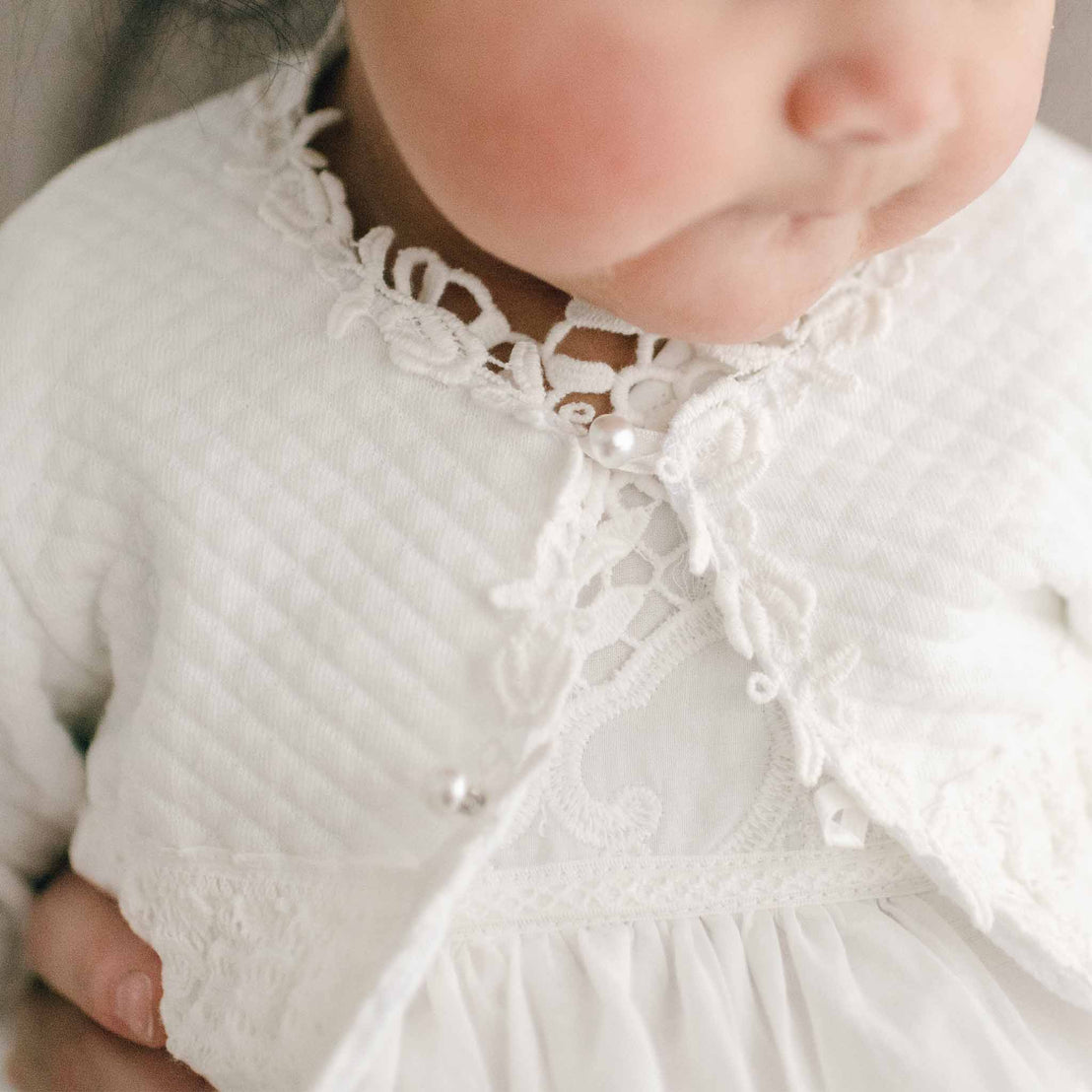 Detail of a baby wearing a Lily Quilted Cotton Sweater. The photo showcases the bodice that is accented with ivory rose and vine lace