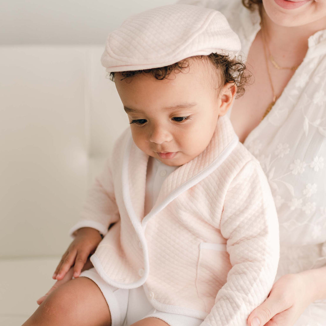 Baby boy sitting on a white couch with his mother and wearing the pink Asher 3-Piece Suit, including a folded collar jacket, white pants and a onesie with buttons (with matching Quilted Newsboy Cap).