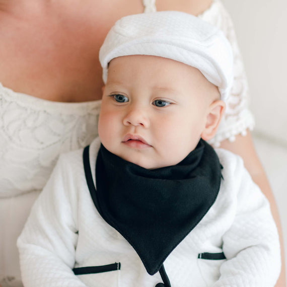 A baby wearing the white James 3-Piece Suit with matching newsboy cap and the James Bandana Bib. The bib is made out of black French Terry and features a "bandana" design. The baby is sitting in the lap of his mother dressed in a white lace garment. The baby has a calm expression. The background is light-colored and out of focus.