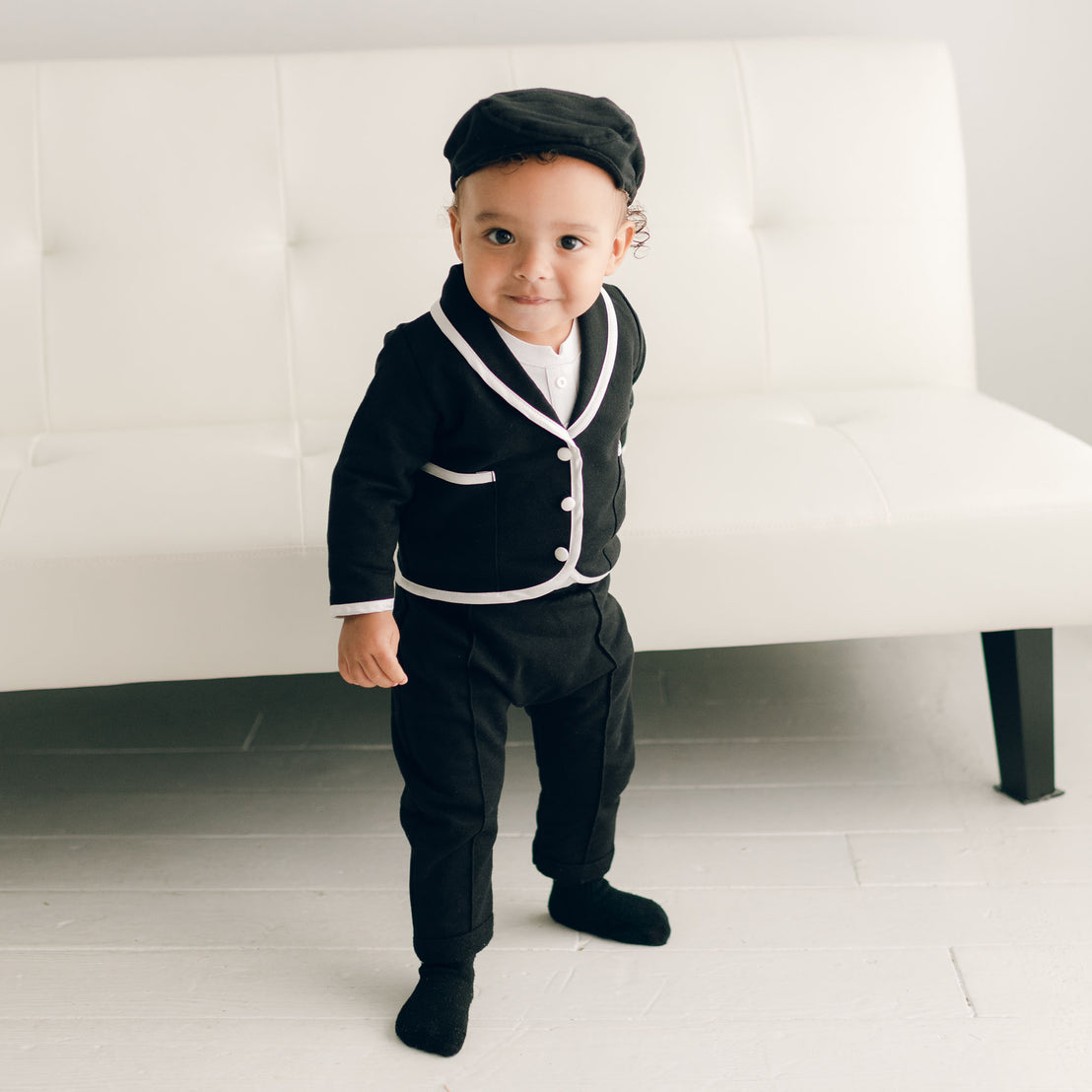Baby boy standing up and wearing the black James 3-Piece Suit, including the jacket, pants, and onesie (with matching Newsboy Cap).