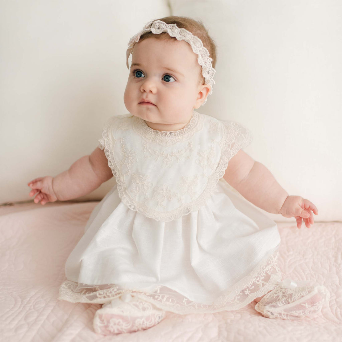 A baby with blue eyes and chubby cheeks is seated on a light pink quilt. The baby is wearing a white lace dress, a Jessica Cotton Bib, and a champagne lace headband. The background is soft and light-colored with white pillows.