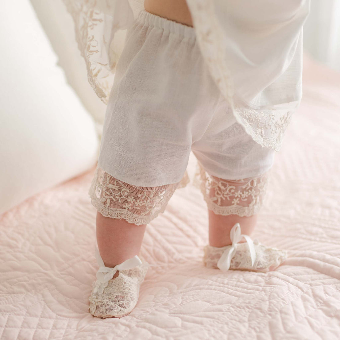 A baby's legs are adorned in the Jessica Linen Dress & Bloomers, featuring white lace-trimmed shorts and matching lace shoes with ribbons, adding a vintage touch. This delicate ensemble complements the soft, pink quilted surface on which the baby stands.