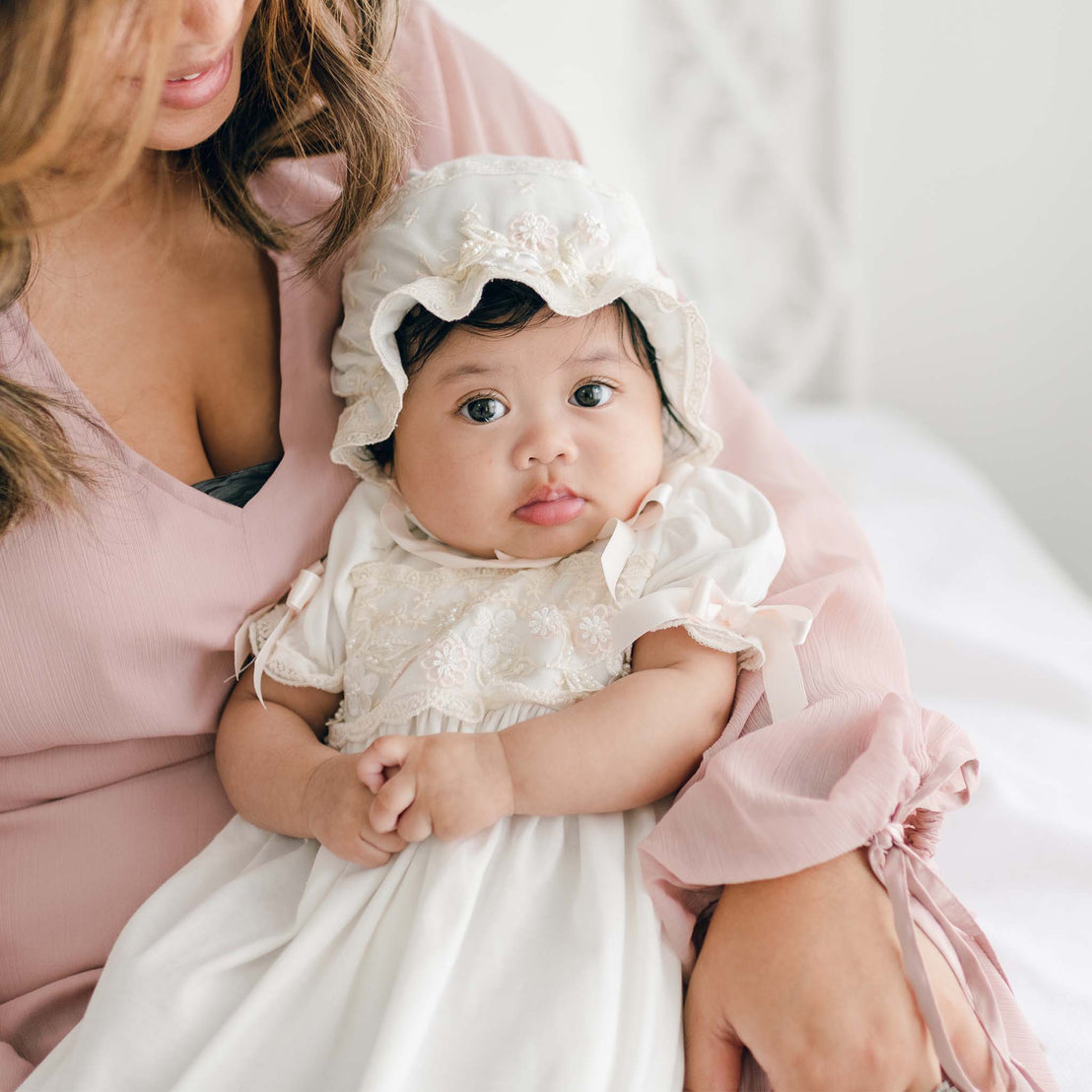 A woman cradles a baby dressed in the Jessica Newborn Gown & Bonnet Set. The woman wears a pink blouse. The baby looks at the camera with big eyes, while the woman gazes affectionately at the baby.