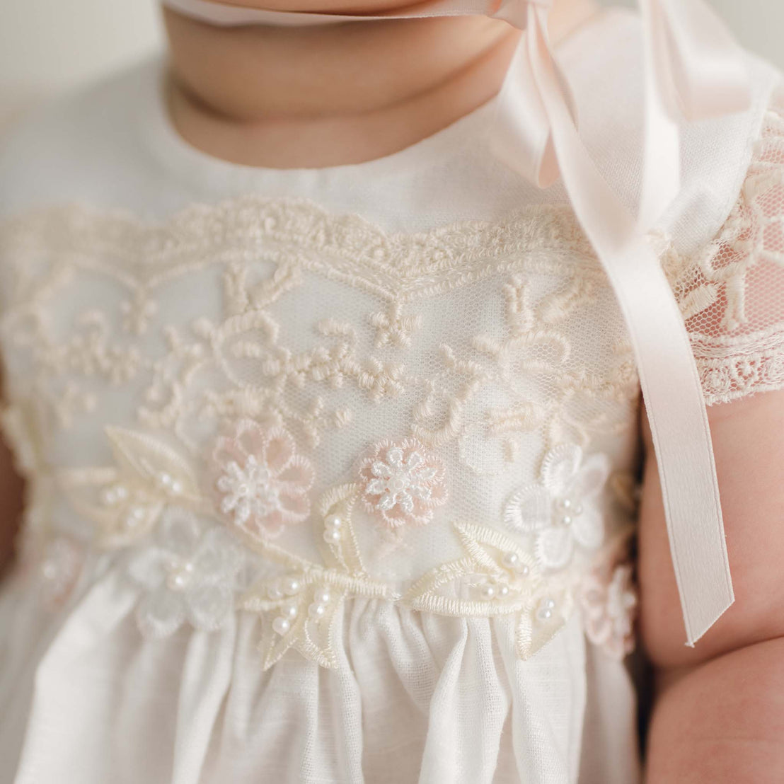 Close-up of a baby wearing the delicate Jessica Linen Dress & Bloomers, featuring a white lace design embellished with floral embroidery and small pearls. The vintage touch is highlighted by lace flutter sleeves, while a satin ribbon tied at the shoulder adds a soft, elegant element—perfect for a Christening gown.