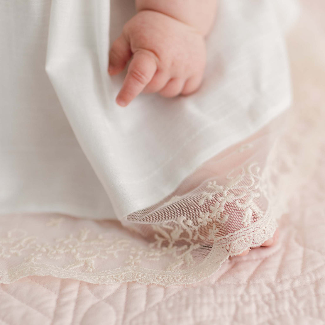 A baby's hand gently grasps the edge of the delicate Jessica Linen Dress & Bloomers set. The fabric, featuring intricate embroidery and lace flutter sleeves, rests on a soft, quilted pink surface.