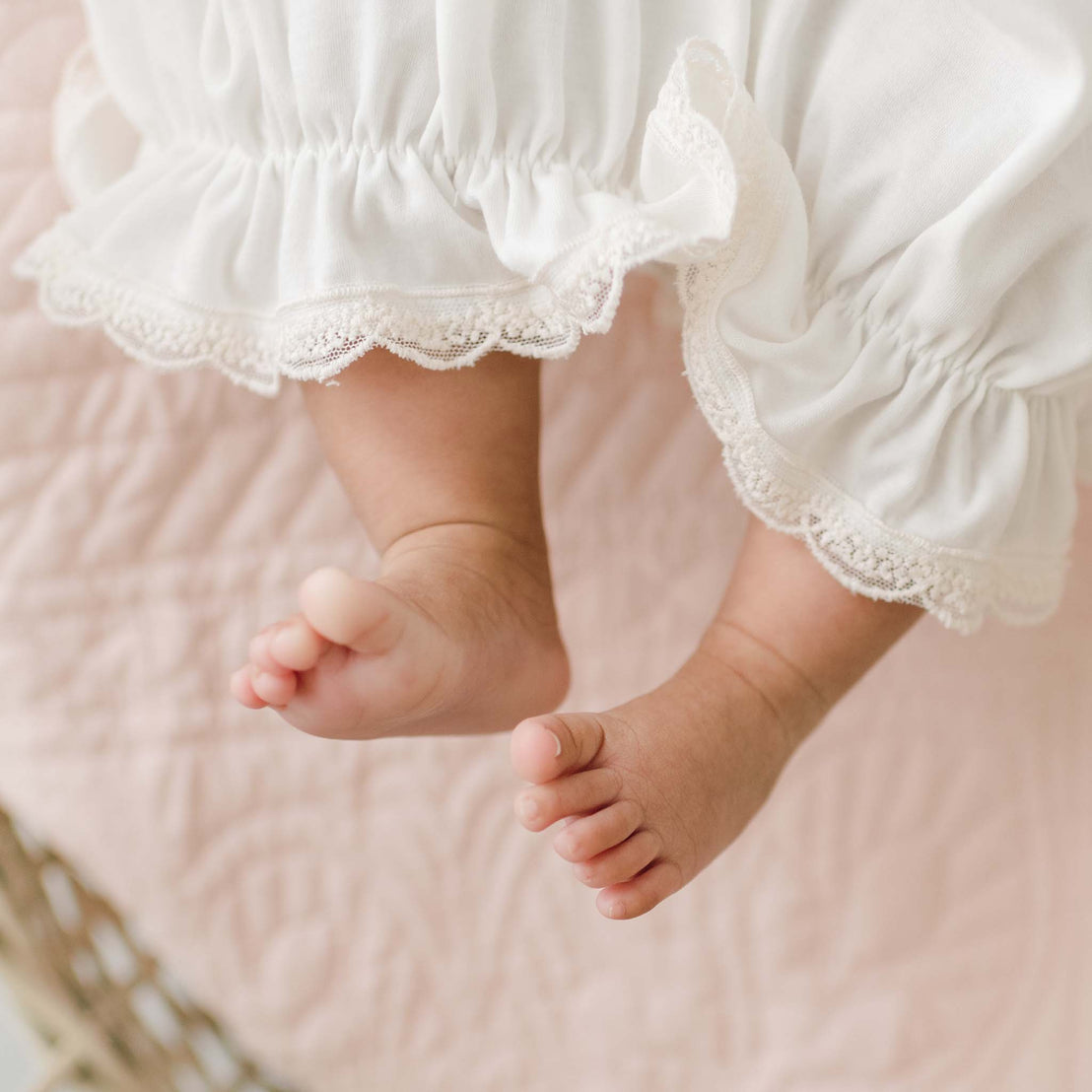 Close-up of an infant's feet, partially covered by the ruffled hem of a Jessica Newborn Gown & Bonnet Set. The baby is lying on a soft, light pink quilt, and the gentle light suggests a calm, cozy setting.