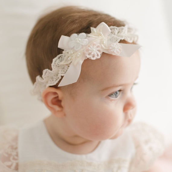 A baby in a Jessica Headband and a handmade headband with pastel floral appliqué sits on a white surface, gazing to the right. The soft lighting enhances the serene and delicate atmosphere of the scene.