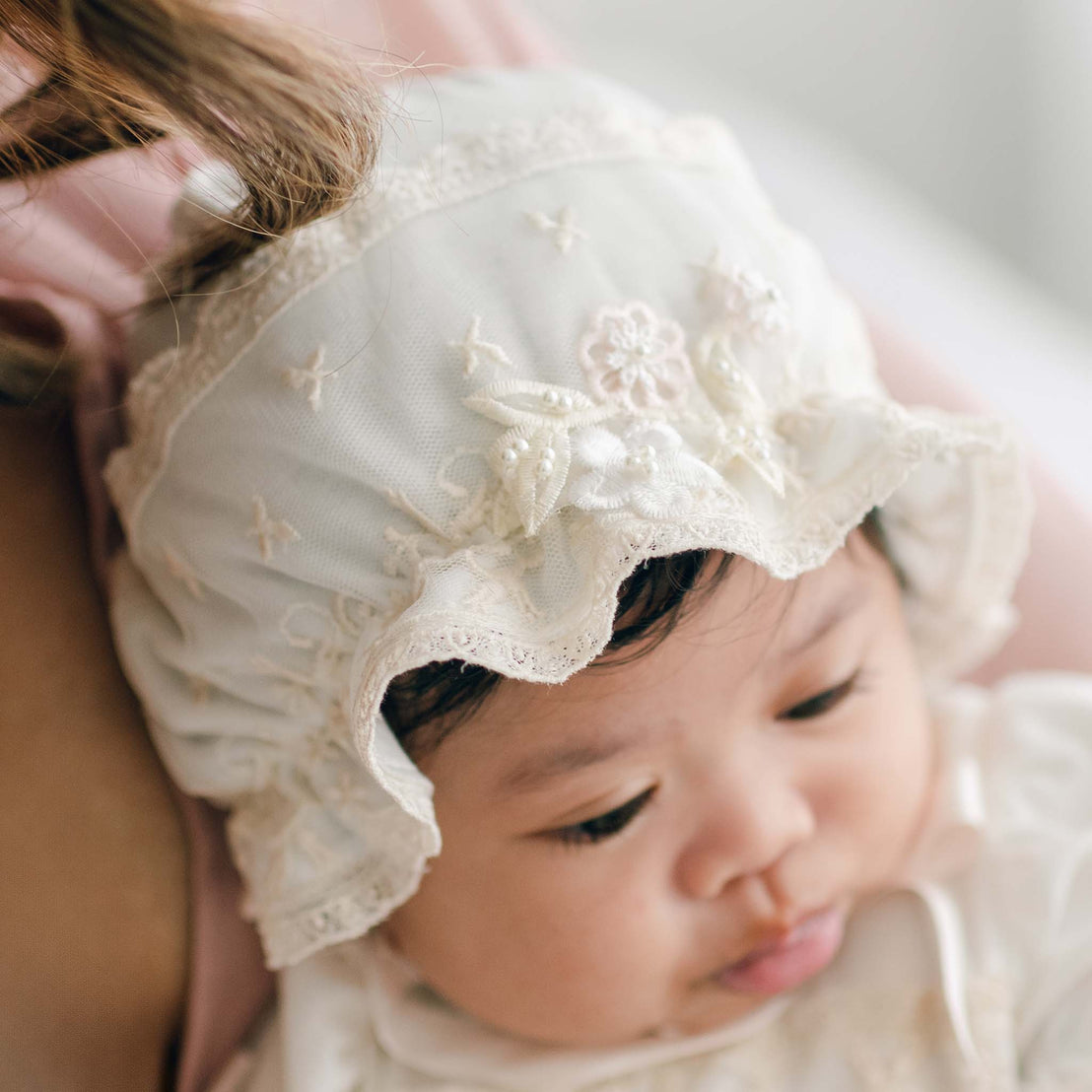 A close-up of a baby wearing an ornate white bonnet with floral embroidery and lace trim. The infant, dressed in a delicate Jessica Newborn Gown & Bonnet Set, is leaning against someone and gazing downward. The background is softly blurred.