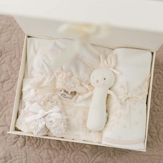 A neatly arranged gift box contains delicate baby items, including a handmade baby gown with floral applique, a pair of lacy booties tied with white ribbons, a soft white rabbit plush toy with pink ears, and a cozy white blanket adorned with subtle floral patterns. Perfect for a newborn girl gift is the Jessica Newborn Gift Set - Save 10%.