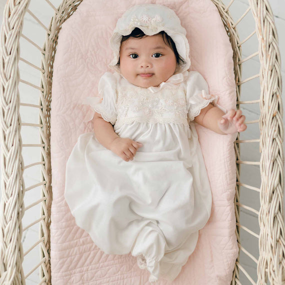 A baby dressed in a Jessica Newborn Gown & Bonnet Set is lying on a pink quilted blanket inside a wicker basket. The baby girl, in her lovely newborn outfit, is looking at the camera with a slight smile and one hand slightly raised.