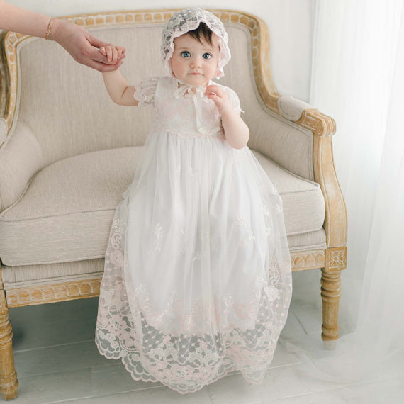 A baby in a white Joli Christening Gown and bonnet sits on an elegant couch, with a hand holding theirs, against a soft-hued backdrop.
