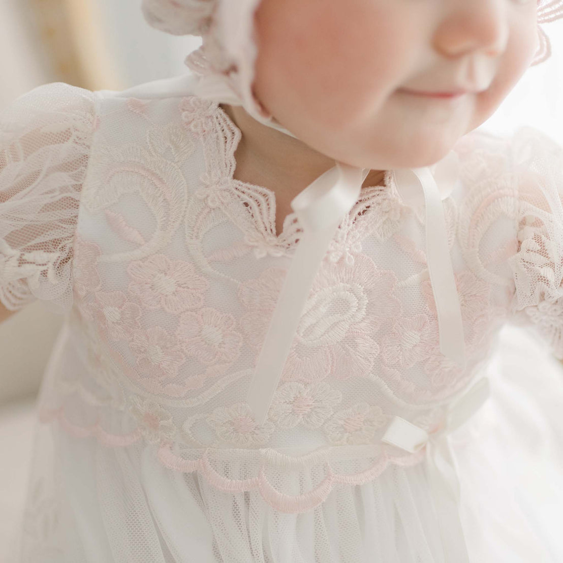 Close-up of a toddler girl in a delicate white Joli Christening Gown with pink trim and ribbon, focusing on the intricate fabric and soft texture around her upper body and neck.
