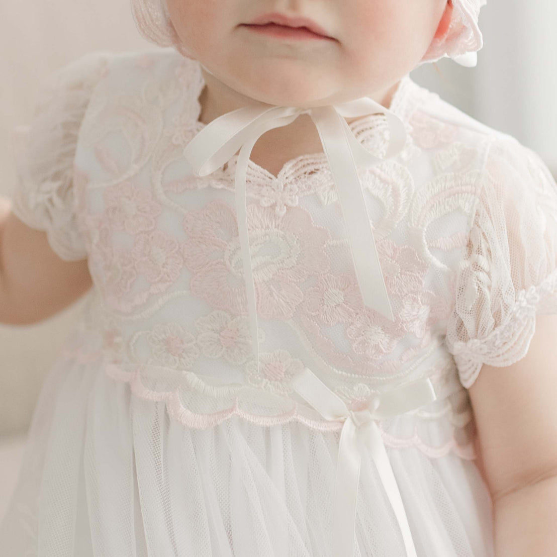 A close-up of a baby in a Joli Christening Gown with short sleeves and a ribbon tied in a bow at the waist, focusing on the detailed fabric and bow.