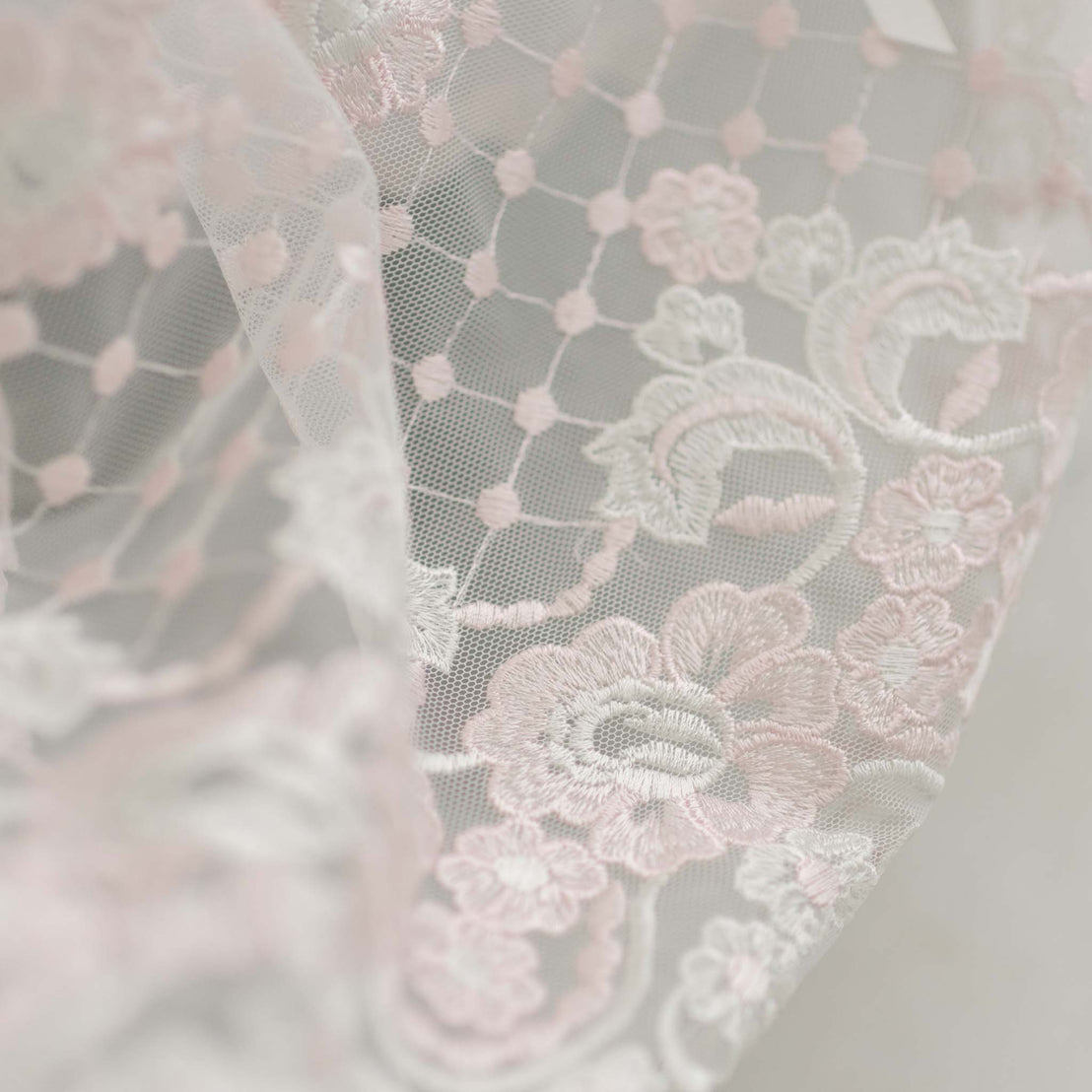 Close-up of a delicate white lace Joli Christening Gown with pink floral embroidery and mesh background, illustrating intricate textile details in the embroidered netting.