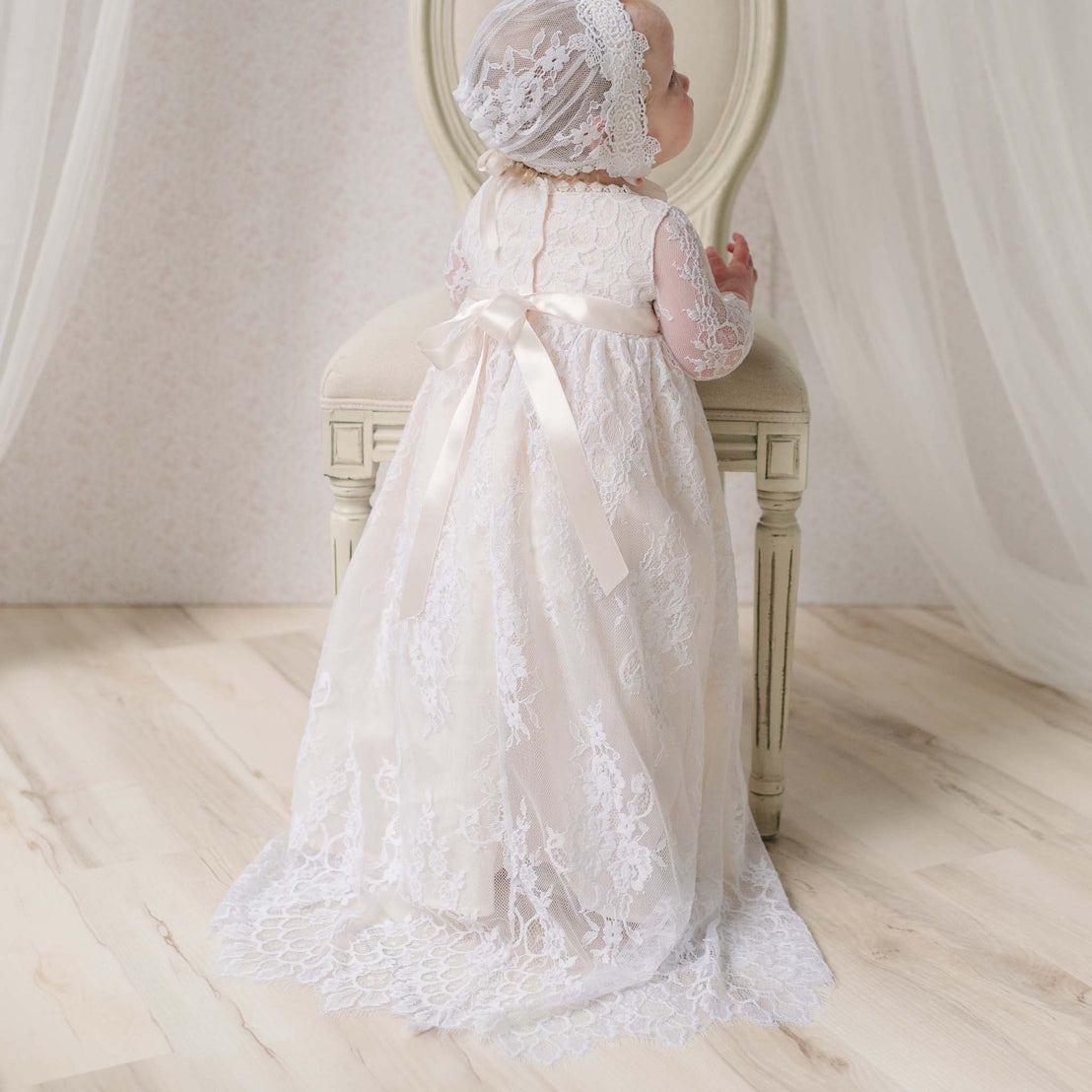 Baby in a white and pink lace gown standing with a chair on a soft, neutral background.