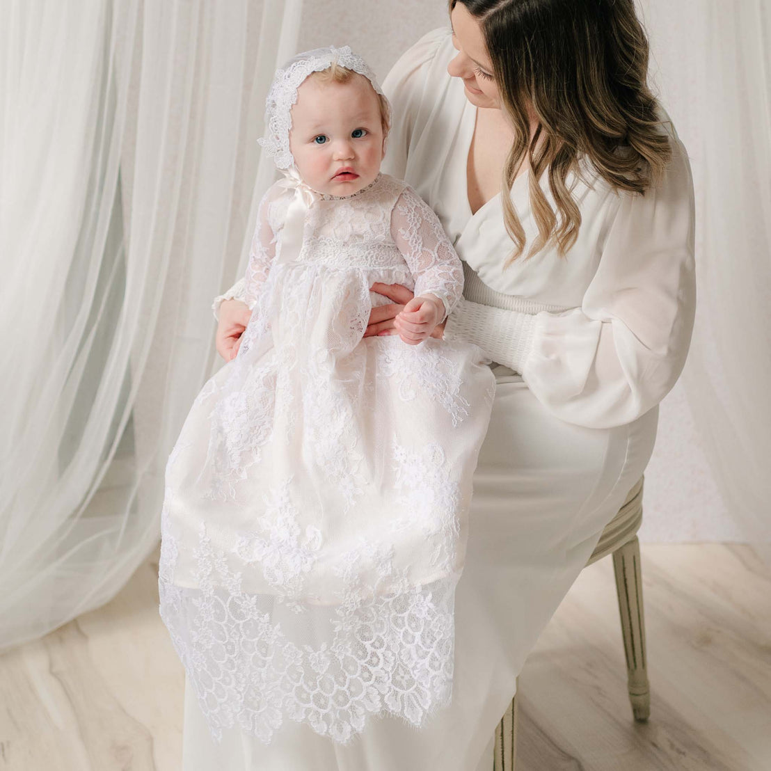 Mom sitting holding a baby dressed in a white lace gown and bonnet against a white curtain background